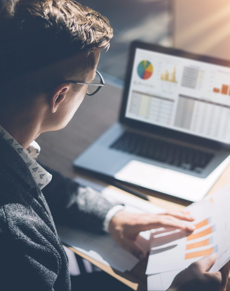 Young finance market analyst in eyeglasses working at sunny office on laptop while sitting at wooden table.Businessman analyze document in his hands.Graphs and diagramm on notebook screen.Vertical